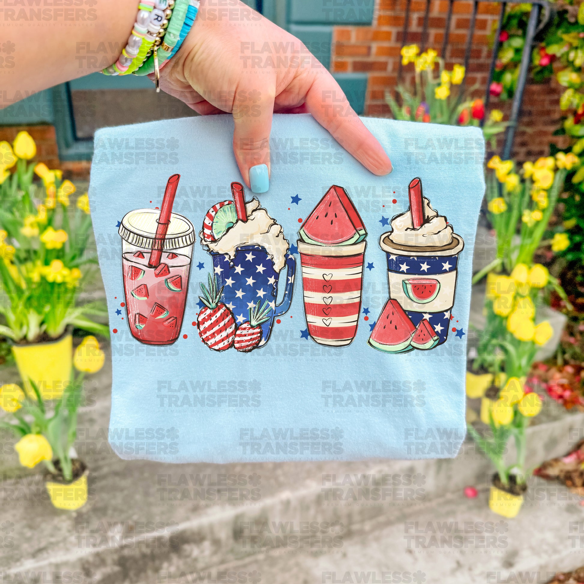 A hand holding a blue shirt with a patriotic design featuring four colorful drinks, including a red and white striped drink, a watermelon drink, and two iced coffee drinks.