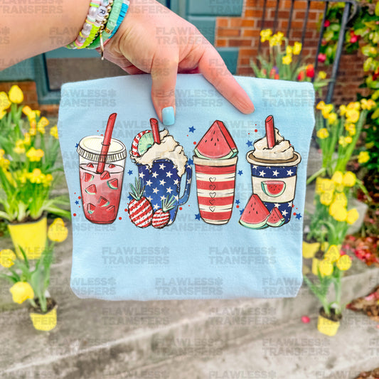 A hand holding a blue shirt with a patriotic design featuring four colorful drinks, including a red and white striped drink, a watermelon drink, and two iced coffee drinks.